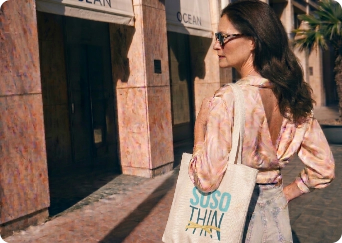 Woman walking on a sunny street, carrying a beige tote bag with turquoise text that reads 'SUSO THIN' over her shoulder.