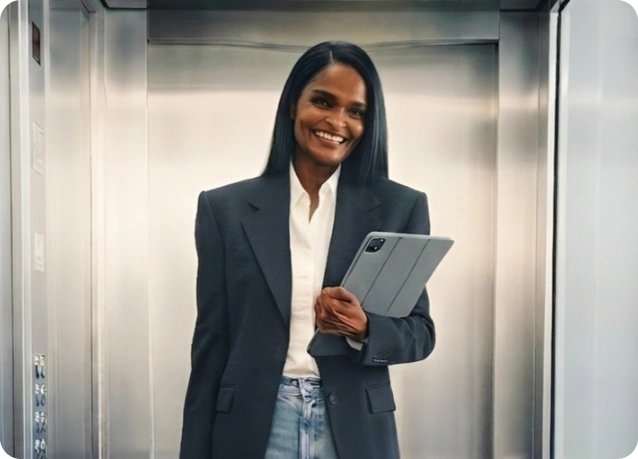 Woman in a dark blazer and white shirt smiles while holding a tablet inside an elevator, ready for a meeting or presentation.