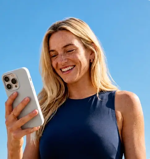 Woman with long blonde hair in a navy sleeveless top smiling while looking at a smartphone against a blue sky.